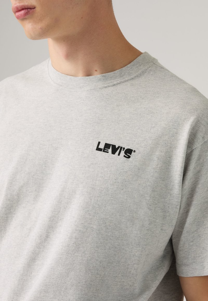 Mann trägt hellgraues Levi's T-Shirt mit schwarzem Logo auf der linken Brust, sichtbar vom Hals bis zur Mitte des Torsos vor einfarbigem Hintergrund.
