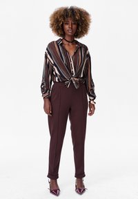 Striped blouse in brown, cream, and navy, knotted at the waist. High-waisted brown trousers with a smooth texture and rolled cuffs. Burgundy heels.