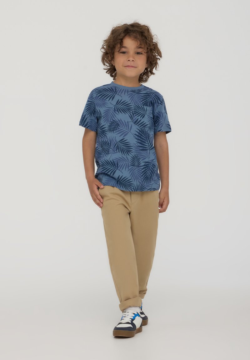 T-shirt azzurro a maniche corte con motivo di foglie di palma, abbinato a pantaloni beige e scarpe blu navy con dettagli bianchi e suola marrone.