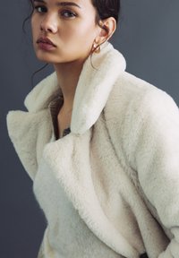 Kobieta w białym futerku z ekologicznego materiału i złotych kolczykach-kołach, patrząca lekko na bok na tle jednolitego ciemnego tła.