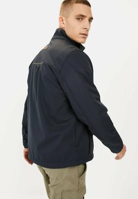 Marineblaue Jacke aus wasserabweisendem Material, mit hohem Kragen, Reißverschlusstaschen und einem horizontalen Akzentstreifen auf dem Rücken.