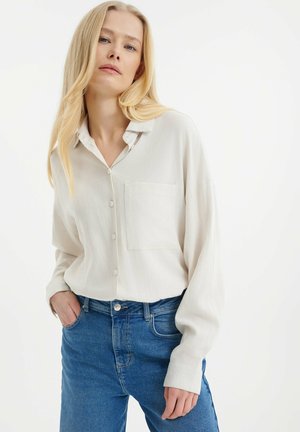 Helles beige Button-Down-Hemd mit Brusttasche; lange Ärmel, lockere Passform. Kombiniert mit blauen Jeans, glatte Textur, dezenter Stoffglanz.