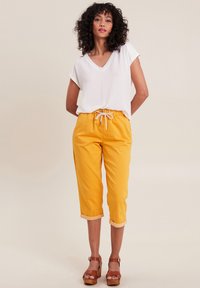 Witte blouse met korte mouwen en een v-hals, gecombineerd met felgele cropped broeken met een trekkoord in de taille en omslagmanchetten. Bruine platform sandalen.