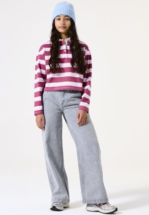 Gestreiftes, pinkes und weißes Crop-Polo, kombiniert mit hellgrauen weit geschnittenen Jeans. Das Model trägt eine blaue Strickmütze und mehrfarbige Turnschuhe.