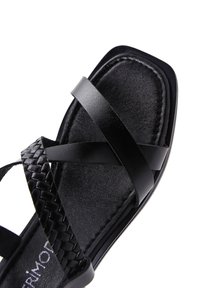 Sándalo de piel negra con suela plana, diseño de tiras trenzadas y múltiples tiras cruzadas. La textura es suave con un ligero brillo.