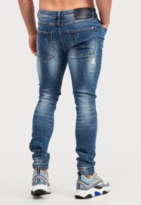 Tunna blå jeans med slitna detaljer, fem fickor och en mörk midjebandsetikett. Bärs med sportiga grå och blå sneakers.