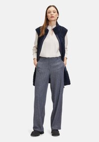Marine fleece-gevoerde vest met hoge kraag, witte blouse en broek met houndstooth-patroon. Zwarte instapschoenen maken de outfit compleet.