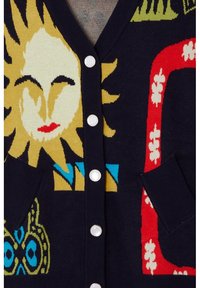 Färgglad stickad cardigan med ett mönster av sol och fjärilar, marinblå bakgrund, vita knappar och två fickor. Texturerad tyg med djärva designelement.