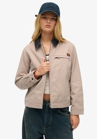 Beige Jacke mit schwarzem Kragen, Frontreißverschluss und einer Brusttasche. Kombiniert mit einer dunkelblauen Mütze. Getragen über einem weißen Oberteil und blauen Jeans.