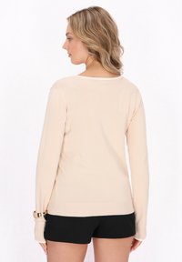 Pulover bej cu mânecă lungă și guler rotund, cu tiv și accente la mansete dense. Textură netedă; purtat cu pantaloni scurți negri, evidențiind un aspect casual.