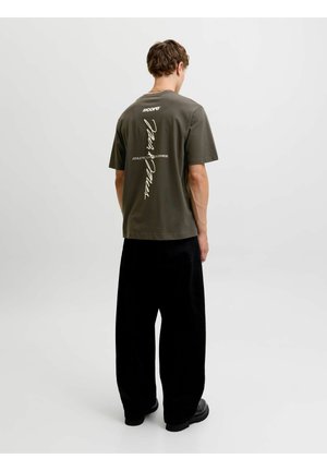 Jeune homme debout, de dos, portant un t-shirt vert olive avec un texte blanc vertical et un pantalon noir ample sur un fond uni.