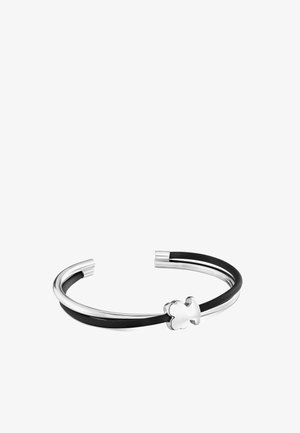 Pulsera de puño abierta en plata y negro con una pequeña decoración de trébol de cuatro hojas en plata sobre fondo blanco.