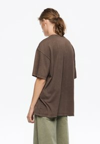 Svetlo rjav oversized T-shirt s okroglim izrezom, kratkimi rokavi in vidnimi šivi, v kombinaciji s svetlo zelenimi hlačami.