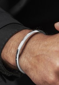 Sølv armring med et elegant, buet design, med en polert overflate og subtil tekstur, båret på et håndledd med lys hår.