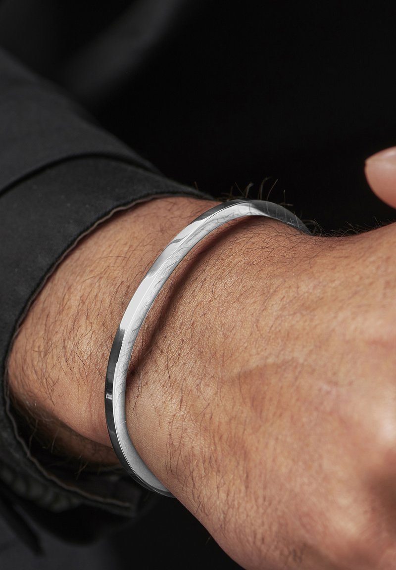 Sølv armring med et elegant, buet design, med en polert overflate og subtil tekstur, båret på et håndledd med lys hår.