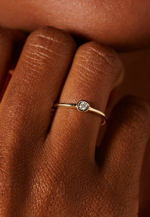 Gouden ring met een enkele heldere ronde edelsteen, gedragen aan de middelvinger van een donkergekleurde hand tegen een wazige achtergrond.