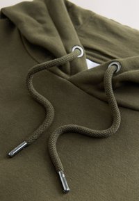 Gros plan sur la capuche d'un sweat à capuche vert olive avec des cordons épais passés à travers des œillets métalliques et des embouts métalliques aux extrémités.