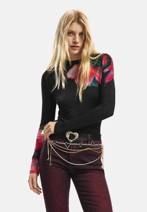 Langærmet sort top med farverigt blomsterprint på ærmerne; kombineret med mørkerøde jeans og en metallic bæltespænde i form af et hjerte.