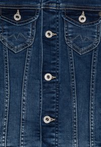 Vaatteet ovat denimliivi syvän sinisen värin, jossa on kaksi rintataskua, hopeiset napit ja erottuvat tikkaus yksityiskohdat.