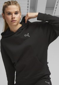 Čierna mikina s kapucňou vyrobená z bavlneného zmesi, s klokankovou vreckom, rebrovanými manžetami a vyšívaným logom Puma v bielej farbe. Hladký povrch.