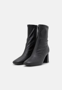 Bottes de cheville en cuir noir avec une fermeture éclair à l'avant, pointes effilées et un talon bloc, présentant une texture plissée et des détails de couture visibles.