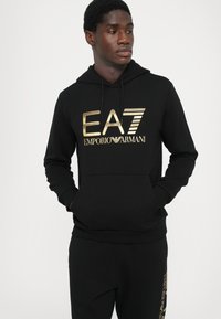 Fekete kapucnis pulóver puha anyagból, aranyszínű "EA7 EMPORIO ARMANI" logóval a mellkason és állítható zsinórokkal.