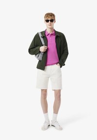 Modell bär en grön jacka, rosa polotröja och vita shorts. Accessoriserad med en grå axelväska och solglasögon. Skorna är vita med gröna accenter.