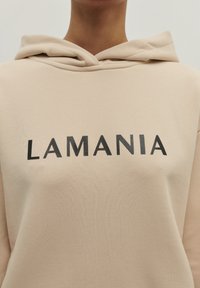 Bežová mikina s klokankou, na prednej strane s tučnými čiernymi nápisom "LAMANIA", jemná textúra a kapucňa so šnúrkou.