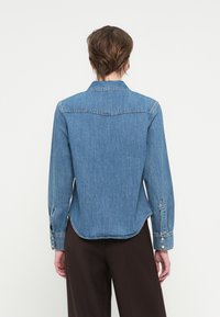 Jeansowa koszula z długimi rękawami, klasycznym kołnierzykiem i zaokrąglonym dołem. Jasnoniebieski kolor, z guzikami przy mankietach i subtelnie zdobionymi szwami.