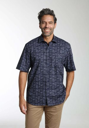 Homme souriant portant une chemise à manches courtes bleu marine à motifs et un pantalon marron clair, debout devant un fond clair uni.