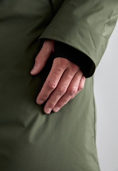 Olive green puffer jacket sleeve with smooth texture, featuring a black inner cuff and a hand resting on the fabric, displaying creases.