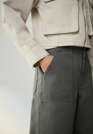 Személy bézs színű dzsekit visel, amelyen fedeles zseb található, és sötétszürke nadrágot, kezét az elülső zsebben tartja, világos háttér előtt.