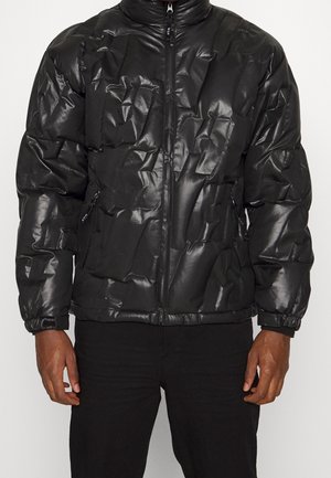 Mężczyzna w błyszczącej czarnej pikowanej kurtce puchowej z przednim suwakiem i zapinanymi na zamek kieszeniami po bokach, w zestawieniu z czarnymi spodniami