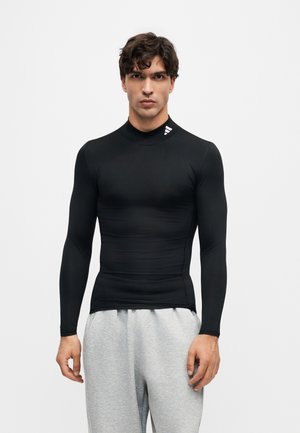 Mann trägt ein eng anliegendes schwarzes langärmeliges Sportoberteil mit kleinem weißen Logo nahe dem Kragen und hellgraue Jogginghose, steht vor einfachem Hintergrund.