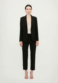 Femeie îmbrăcată în sacou și pantaloni negri, cu un top din dantelă albă și sandale cu toc nude, stând în fața unui fundal alb simplu.