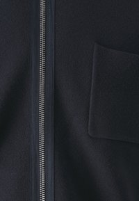 Gros plan d'une veste en tissu foncé montrant une fermeture éclair argentée et une poche carrée sur la poitrine côté droit.