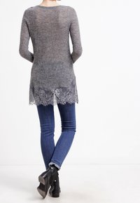 Pull en tricot gris avec une bordure en dentelle à l'ourlet, associé à un jean slim bleu et des bottines noires, mettant en valeur des détails en dentelle complexes.