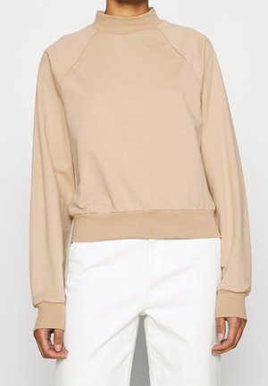 Bluza taupe z długimi rękawami, wysokim kołnierzem i prążkowanymi mankietami, zestawiona z białymi spodniami na stojącej osobie na jednolitym tle.