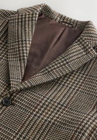 Brūns un pelēks kvadrātveida žakete ar nogriezto apkakli, ar gludu brūnu oderi un vienu melnu pogu priekšā.