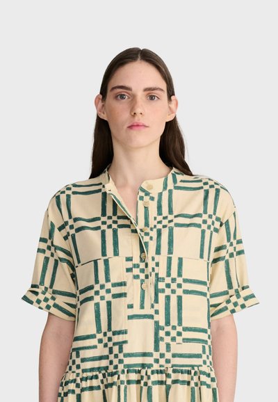 Femme aux longs cheveux foncés portant une robe crème avec un motif géométrique vert en grille et des manches courtes, sur un fond clair uni.
