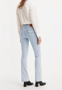 Světle modré džíny s rozšířenými nohavicemi a surovým lemem, vysokým pasem a detaily s kapsami, spárované s bílou košilí s dlouhým rukávem a černými botami na podpatku.