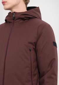 Bordo spalvos kapisonas, pagamintas iš lygaus, vandeniui atsparaus medžiagos, su priekyje užtrauktuku ir užtrauktuku užsegama kišenė kairėje pusėje. Juodas logotipas ant rankovės.