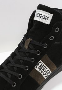 Baskets montantes noires en daim avec des accents en tissu gris et marron, comprenant une étiquette de marque sur la languette et des lacets noirs.