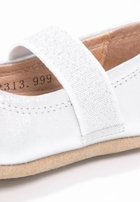 Hvit lærsko med en strukturert elastisk stropp, rund tå og en myk beige semsket såle. Sømmer langs kantene er synlige.