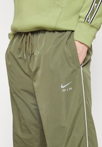 Olivově zelené Nike tepláky vyrobené z lehkého materiálu, s elastickým pasem, šňůrkou a bílými bočními detaily s logem.