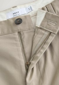 Pantalon beige avec fermeture éclair ouverte, bouton et l'étiquette "NEXT Barrel Fit" indiquant la taille 34R à l'intérieur de la ceinture.
