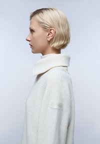 Witte coltrui met een ribbeltextuur, oversized kraag en subtiele merkaanduiding op de mouw. Gladde stof en een losse pasvorm.