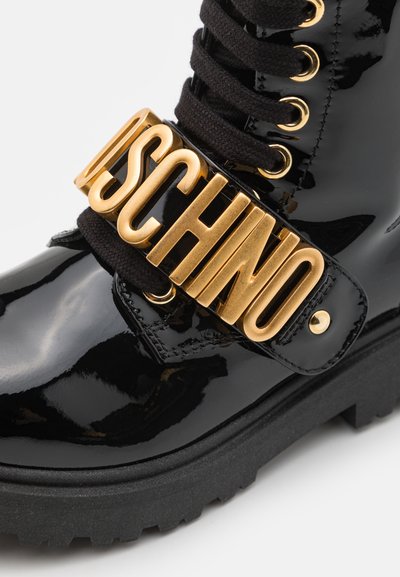 Bottine en cuir verni noir avec un matériel doré en forme de logo sur une sangle proéminente, des lacets noirs épais et une semelle épaisse.