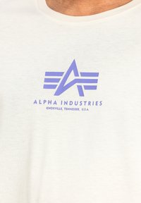 Kerge beež T-särk, mis on valmistatud pehmest kangast. Särgile on trükitud violetne logo, millel on stiliseeritud tähed ja triibud. Tekst sisaldab "ALPHA INDUSTRIES" ja asukohta.