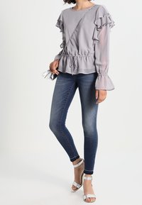 Blusa grigia chiara con maniche arricciate, vita svasata, abbinata a jeans skinny blu scuro e sandali con cinturini argentati, mentre tiene una borsa piccola.